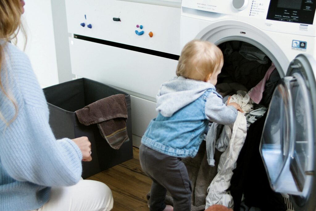 Adorable 1-year-old toddler engaging in learning activities while helping with simple indoor laundry chores alongside mother.
