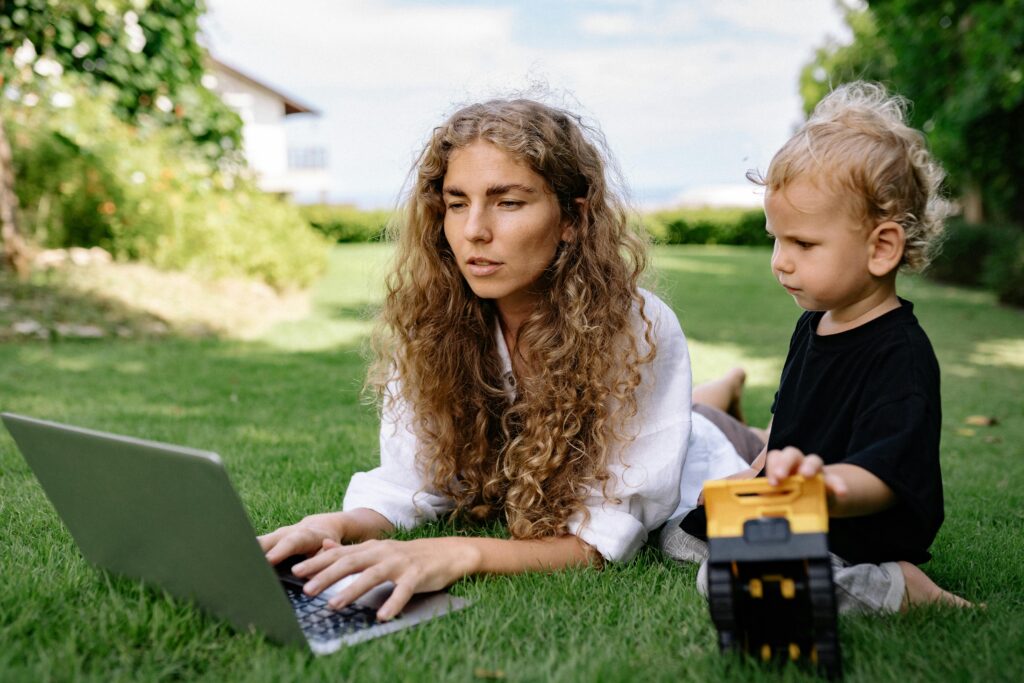 Mother working on a laptop while her toddler plays nearby on the grass enjoying simple outdoor toddler games