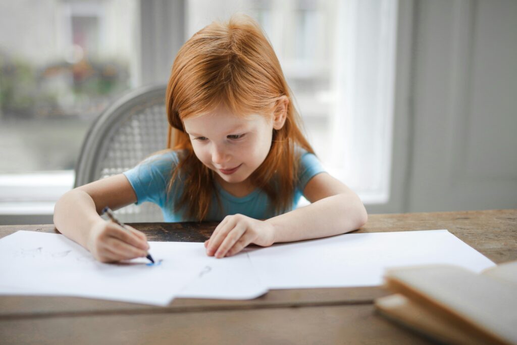 A young girl with red hair focused on drawing at a wooden desk indoors following games and tools for kids.