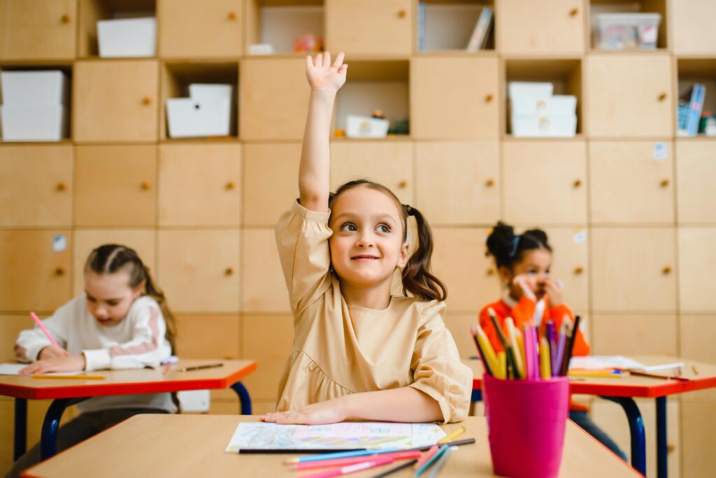 Three children engaged in learning activities inside a classroom, with one raising her hand.